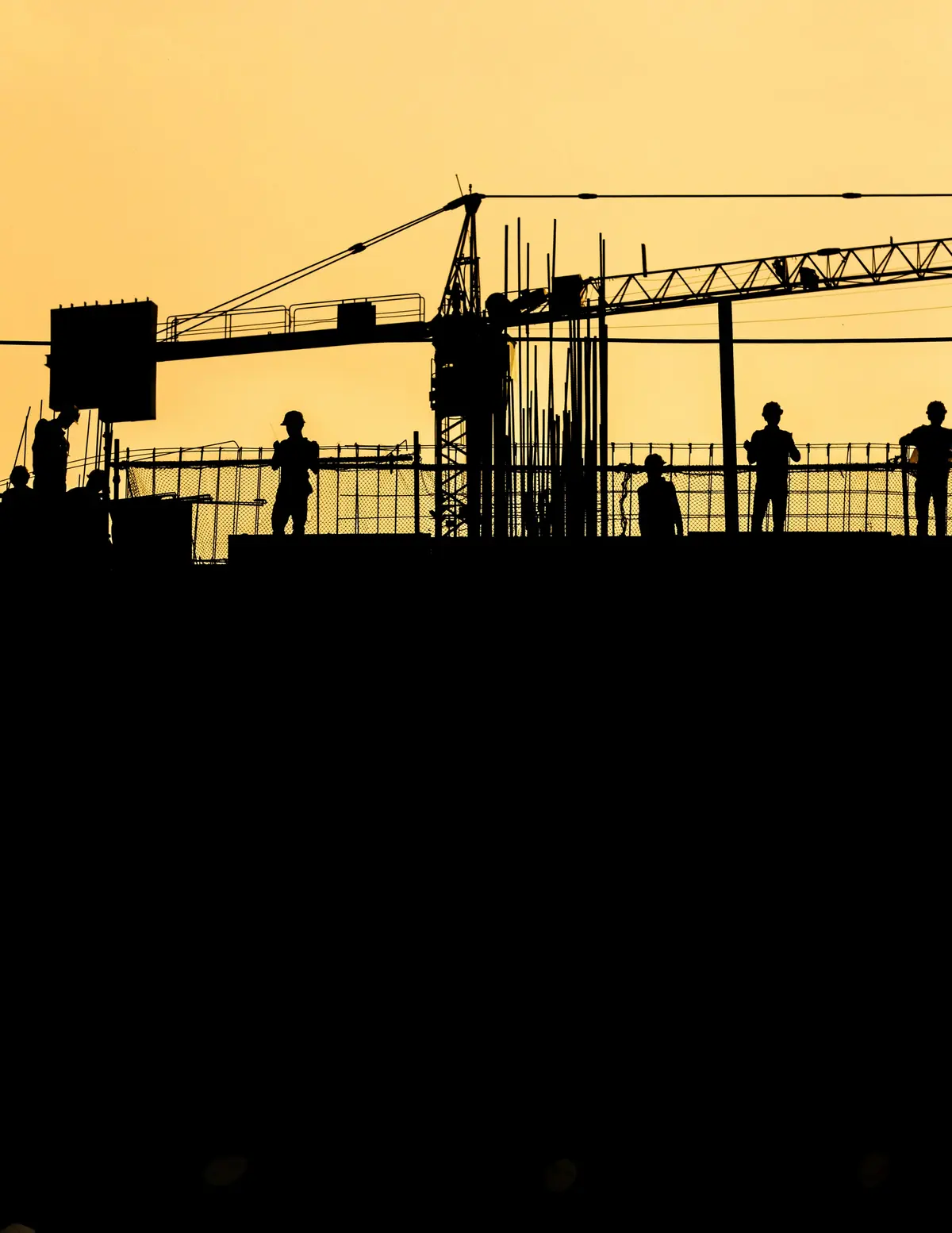 Construction workers in hard hats silhouetted on a rooftop site at sunset, with a crane behind them.